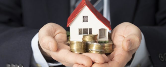 Hands holding a house model surrounded by coins and banknotes, symbolizing real estate investment.