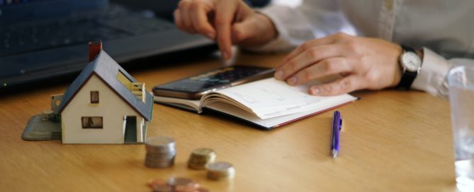 Person calculating finances with a house model and coins on the table.