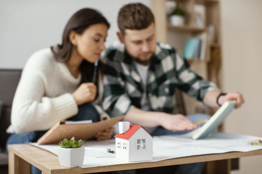 A couple reviewing house plans together at a table with a small house model and a succulent plant in the foreground. 